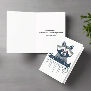 Funny raccoon greeting card open to show message "Here's to us - besties in the same dumpster fire since day one"