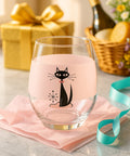 Clear glass with black cat design on pink liquid, placed on a marble surface with gift box and wine bottle in the background.