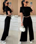 Woman in a black outfit with wide-leg pants, holding a white handbag, in a minimal indoor setting.