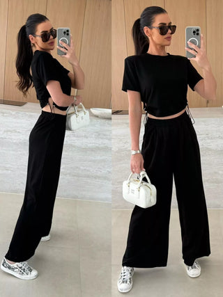 Woman in a black outfit with wide-leg pants, holding a white handbag, in a minimal indoor setting.