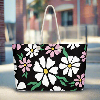 Black oversized tote bag with white and pink daisy flowers and cream rope handles on city sidewalk