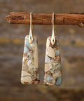 Handcrafted natural stone drop earrings with gold-plated accents, hanging from a rustic wooden branch.