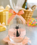 Decorative glass bottle with a black cat design and pink ribbon on a table.